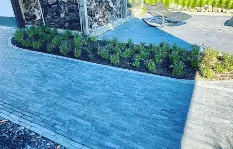 Ein geschwungener Gartenweg aus grauen Pflastersteinen, begrenzt von einem frisch bepflanzten Blumenbeet und Kies, führt zu einer gemütlichen Ruheecke mit einer modernen Schaukelliege neben einem Brennholzunterstand.