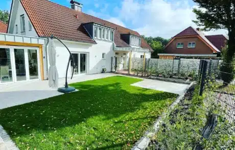 Ein modernes Haus mit weißen Fassaden und roten Ziegeldächern umrahmt einen gepflegten Rasen mit einem großen Schirmständer und angrenzendem Terrassenbereich, umgeben von einem niedrigen Zaun und jungen Heckenpflanzungen.