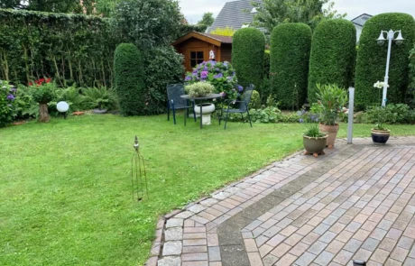 Ein einladender Garten mit einer gemütlichen Sitzgruppe, umgeben von üppigem Grün und farbenfrohen Blumen, im Hintergrund ein charmantes Gartenhaus, eingefasst von akkurat geschnittenen Hecken und einem gepflegten Rasen.