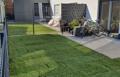 Ein gepflegter Garten mit sattgrünem Rasen neben einem modernen Wohnhaus, begrenzt durch einen Metallzaun. Eine Terrasse mit Gartenmöbeln und ein Fahrrad tragen zum häuslichen Ambiente bei, das durch die ruhige Wohngegend unterstrichen wird.