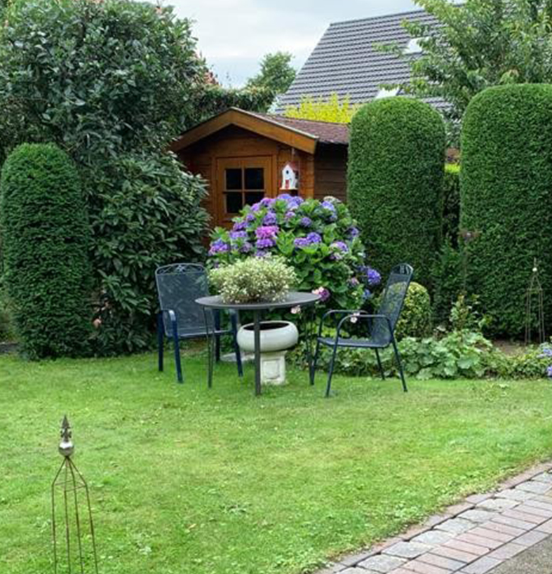 Ein einladender Garten mit einer gemütlichen Sitzgruppe, umgeben von üppigem Grün und farbenfrohen Blumen, im Hintergrund ein charmantes Gartenhaus, eingefasst von akkurat geschnittenen Hecken und einem gepflegten Rasen.