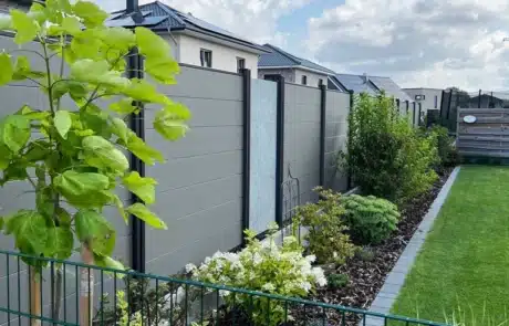 Ein sorgfältig angelegter Garten mit vielfältigen Pflanzen und einem gepflegten Rasen, abgegrenzt durch einen grünen Metallzaun und eine moderne graue Sichtschutzwand, unter einem blauen Himmel mit weißen Wolken.