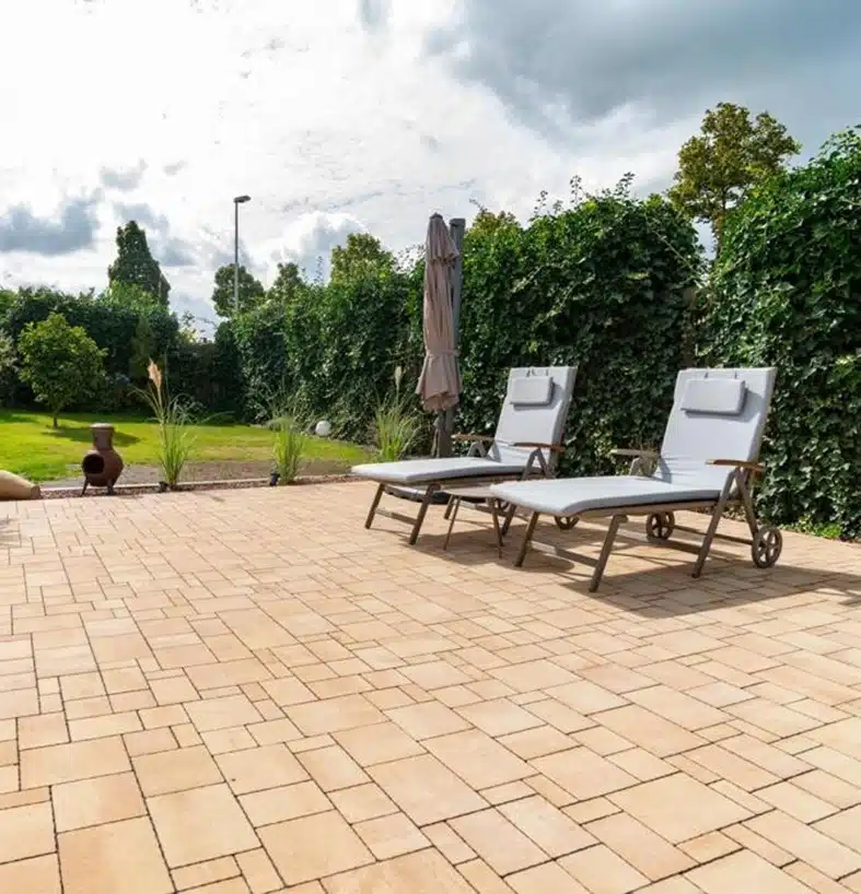 Ein entspannender Außenbereich mit einer großflächigen, sandfarbenen Terrasse, bestückt mit zwei Sonnenliegen und einem Sonnenschirm, umgeben von einer hohen, grünen Hecke und einer gepflegten Gartenlandschaft.